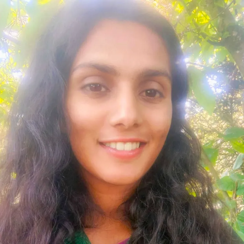 A young woman with long dark curly hair smiling outdoors in a verdant leafy place with dappled sunlight.